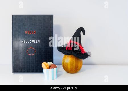 Minimalistisches Halloween Dekor - Brieftafel mit Hello Halloween Botschaft, Hexenhut, dekorativer Kürbis und Tasse mit süßen Makronen auf weißem Hintergrund Stockfoto