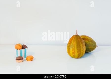 Minimalistisches Halloween-Konzept - Süßigkeiten, Makarons Dessert in einem Papierbecher und dekorative Kürbisse. Schokolade und Kürbismakronen auf weißem Hintergrund Stockfoto