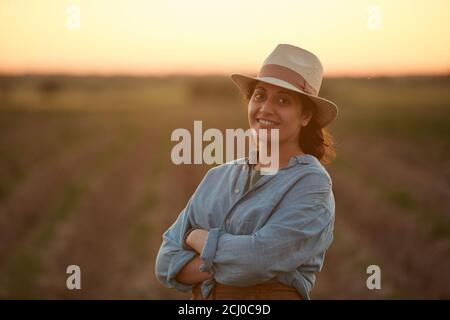 Waist-up-Porträt einer jungen Farmerin, die selbstbewusst mit gekreuzten Armen posiert, während sie bei Sonnenuntergang im Feld steht und die Kamera anlächelt, Raum kopieren Stockfoto