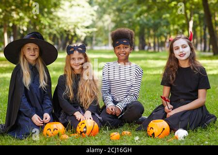 In voller Länge Porträt der multi-ethnischen Gruppe von Kindern tragen Halloween Kostüme und Blick auf die Kamera, während auf grünem Gras sitzen Im Freien Stockfoto