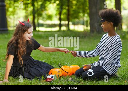 Seitenansicht Porträt von Jungen und Mädchen tragen Halloween-Kostüme Und teilen Süßigkeiten, während auf grünem Gras im Freien sitzen Stockfoto