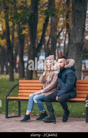 Ein Mann und ein Mädchen ruhen auf einer Bank in einem Herbstpark. Ein liebevolles Paar in Jacken sitzt auf einer Bank im Hauptpark Stockfoto