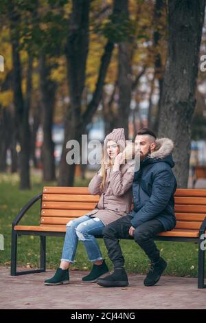 Ein Mann und ein Mädchen ruhen auf einer Bank in einem Herbstpark. Ein liebevolles Paar in Jacken sitzt auf einer Bank im Hauptpark Stockfoto