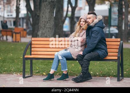 Ein Mann und ein Mädchen ruhen auf einer Bank in einem Herbstpark. Ein liebevolles Paar in Jacken sitzt auf einer Bank im Hauptpark Stockfoto