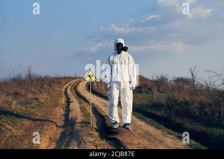 Ökologe in weißem Overall, Gasmaske und Handschuhe stehen auf Dorfstraße auf dem Feld neben Strahlungszeichen, an schönen sonnigen Tag unter blauem Himmel. Konzept von Ökologie, Gefahr und Strahlengefahr. Stockfoto