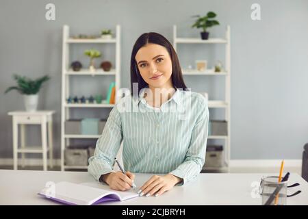 Lächelnde Frau beim Lernen, beim Betrachte der Kamera und beim Aufschreiben von Notizen im Notizbuch während des Online-Unterrichts Stockfoto