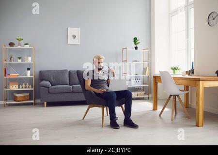 Lächelnder Mann sitzt auf einem Stuhl mit Laptop und arbeitet online Zu Hause Stockfoto