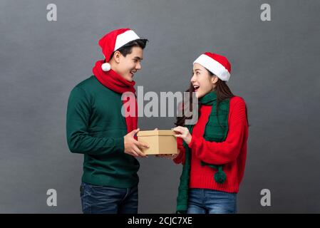 Schöner asiatischer Mann in Weihnachtskleidung überraschend seine Freundin mit Geschenk in einer Schachtel auf grauem isoliertem Studiohintergrund Stockfoto