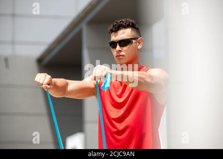 Handsome Sport Mann tun Schulter vorne erhöhen Übung mit Widerstand Band im Freien in der Sonne Stockfoto