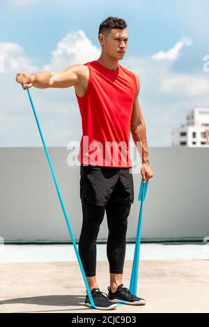 Handsome Sport Mann tun Schulter lateral erhöhen Übung mit Widerstand Band im Freien auf dem Dach in der Sonne Stockfoto