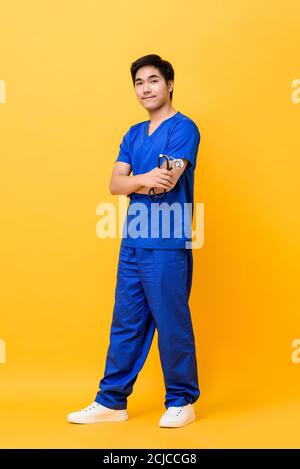 In voller Länge Porträt von lächelnden jungen asiatischen Mann Krankenschwester mit Arme gekreuzt halten Stethoskop in isolierten Studio gelben Hintergrund Stockfoto