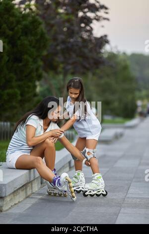 Mutter hilft ihrem kleinen Kind Tochter trägt Rollschuhe im Sommer Park. Stockfoto