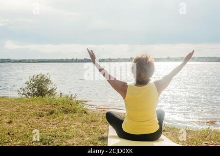 Ältere Frau macht Yoga in der Nähe des Flusses im Sommer. Rückansicht, Hände nach oben. Stockfoto