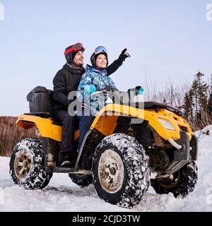 Seitenansicht der fröhlichen jungen Frau und Mann in Helmen sitzen auf All-Terrain-Fahrzeug. Schöner männlicher Reisender zeigt auf etwas und lächelt, während Quad-Bike mit Frau in den Winterbergen fahren. Stockfoto