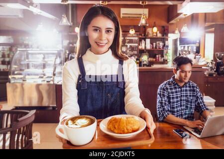 Asian Barista der kleinen Unternehmer serviert eine Tasse Kaffee und Croissants Bäckerei für junge Kunden am Tisch Im Café, Inhaber kleiner Geschäfte Stockfoto