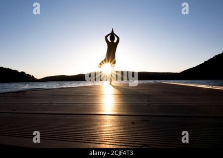 Junges Mädchen tut Yoga Posen auf einem Pier bei Sonnenuntergang. Baumhaltung Stockfoto