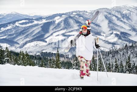 Horizontale Momentaufnahme des jungen Skifahrers in farbenfrohem Kostüm, der entlang verschneiten Hügels geht, seine Skier vor erstaunlichen Bergen im Hintergrund carring. Vorderansicht, Kopierbereich, volle Länge Stockfoto