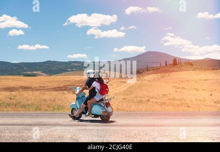 Ein Mann und eine Frau auf einem Roller auf einer Landstraße mit der Sonne scheint in der Toskana. Zypressen, Berge und Villa am Horizont mit blauem Himmel und w Stockfoto