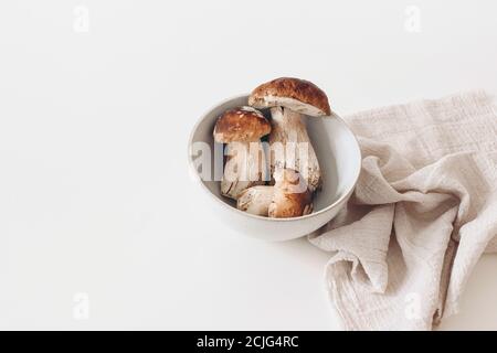 Essbare Boletus edulis, Steinpilze in Keramikschale. Leckere Wildzeps auf weißem Tisch mit beiger Leinenserviette. Leerer Kopierplatz, Minimum Stockfoto