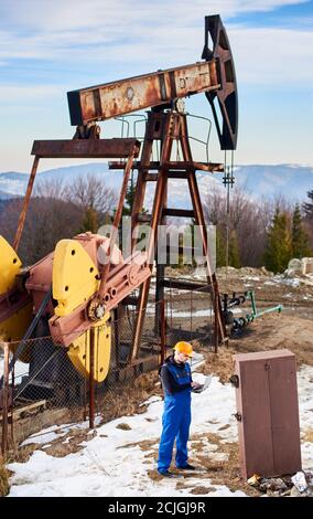 Volle Länge der Petroleum Ingenieur in Arbeitsüberalls und Helm Überprüfung Ölpumpwerk, mit Laptop in der Petroleum-Anlage. Männlicher Bediener, der am Notebook arbeitet, während er in der Nähe der Kippmaschine der Ölpumpe steht. Stockfoto