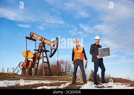 Zwei Männer auf Ölfeld mit Pumpenheber auf Hintergrund. Geschäftsmann hält tragbare Solarpanel, Ölbrunnen Betreiber mit Schraubenschlüssel in der Hand. Konzept der Erdölindustrie und alternative Energiequellen. Stockfoto