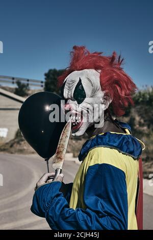 Nahaufnahme eines gruseligen bösen Clowns im Freien, hält einen schwarzen Ballon in der Hand und bringt ihm die Spitze eines großen Messers auf den Mund, während er auf die OBs starrt Stockfoto