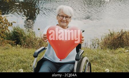 Ältere Frau hält einen herzförmigen Ballon in der Nähe des Flusses. Liebe und Pflege Konzept. Hochwertige Fotos Stockfoto