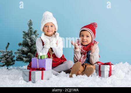 Kinder im Winteroutfit sitzen auf Schnee und gestikulieren in der Nähe Geschenke und Tanne isoliert auf blau Stockfoto