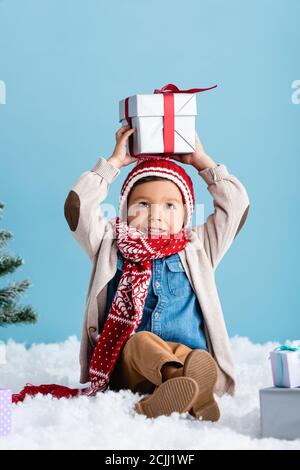 Kind in Hut und Winter-Outfit auf Schnee sitzen und Über dem Kopf halten, isoliert auf blau Stockfoto