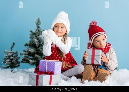 Kinder im Winter Outfit sitzen auf Schnee in der Nähe von Tannen und Präsentiert isoliert auf blau Stockfoto