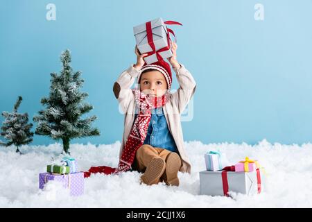 Junge in Hut und Winter-Outfit sitzt auf Schnee und Halten Geschenk über Kopf in der Nähe von weihnachtsbäumen isoliert auf blau Stockfoto