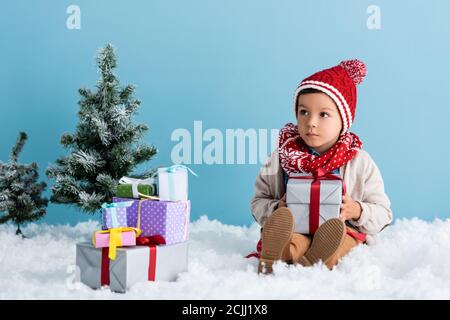 Junge in Hut und Winter-Outfit sitzt auf Schnee in der Nähe Geschenke und weihnachtsbäume isoliert auf blau Stockfoto