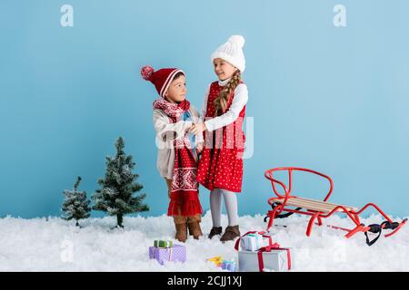 Junge im Hut hält Hände mit Schwester in der Nähe von Kiefern, Geschenke und Schlitten isoliert auf blau Stockfoto