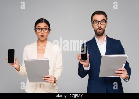 Schockiert interracial Geschäftspartner halten Ordner, während zeigt Handys Mit leerem Bildschirm, der auf grau isoliert ist Stockfoto