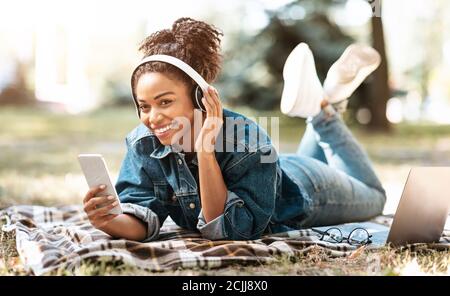 Afrikanische Frau Mit Telefon Tragen Kopfhörer Liegen Auf Plaid Im Freien Stockfoto