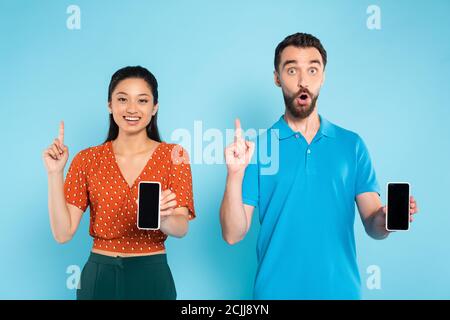 Junge asiatische Frau und aufgeregt Mann zeigt Idee Zeichen, während Halten Sie Smartphones mit leerem Bildschirm auf blau Stockfoto