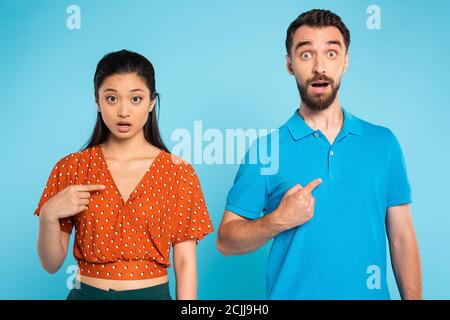 Schockiert asiatische Frau in roten Bluse und bärtigen Mann in polo-T-Shirt zeigt mit den Fingern auf sich selbst auf blau Stockfoto