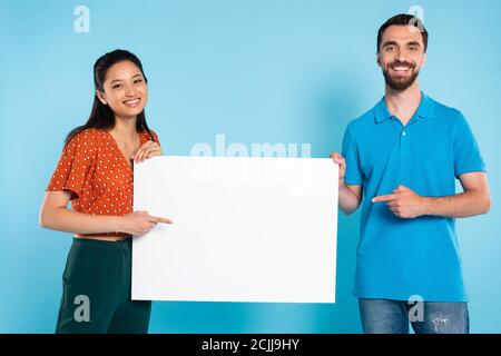 asiatische Frau in roter Bluse und Mann in Polo-T-Shirt Zeigt mit den Fingern auf ein leeres Plakat auf blau Stockfoto