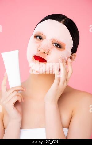 Brunette asiatische Frau berühren Gesichtsmaske während halten Rohr von Kosmetische Creme isoliert auf rosa Stockfoto