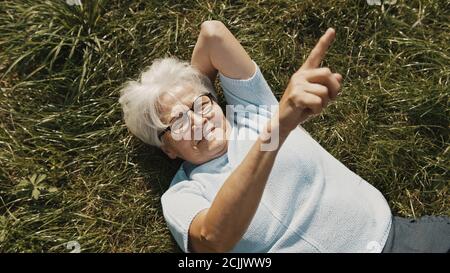 Ältere Frau, die sich auf dem Gras ruht und mit dem Finger in den Himmel zeigt. Hochwertige Fotos Stockfoto