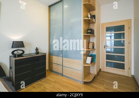 Moderne Einrichtung eines luxuriösen Schlafzimmers. Weiße Wände. Schrank mit Schiebetüren aus Glas und Holzregale mit Büchern. Stockfoto