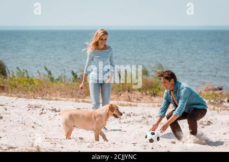 Paar mit Fußball und Golden Retriever spielen am Strand während Wochenende Stockfoto