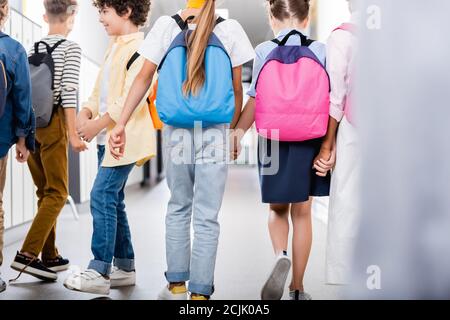 Rückansicht multiethnischer Klassenkameraden, die beim Spaziergang entlang des Schulkorridors die Hände halten, selektiver Fokus Stockfoto