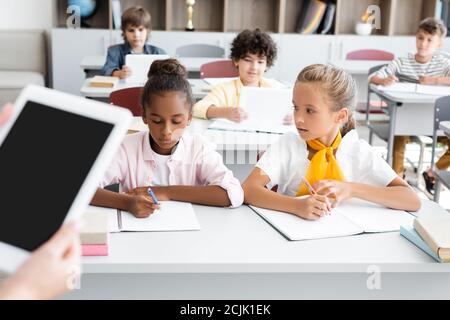 Beschnittene Ansicht des Lehrers mit digitalem Tablet mit leerem Bildschirm In der Nähe multikulturelle Schüler schreiben in Notebooks Stockfoto