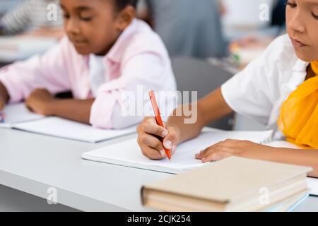 Selektiver Fokus multiethnischer Schülerinnen, die auf Notizbüchern in der Nähe von Büchern schreiben Auf dem Schreibtisch im Klassenzimmer Stockfoto