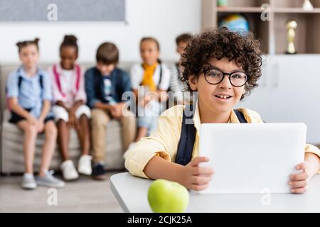 Selektiver Fokus des muslimischen Schuljungen mit digitalen Tablet in der Nähe von Apfel Auf dem Schreibtisch und multiethnische Klassenkameraden im Hintergrund Stockfoto