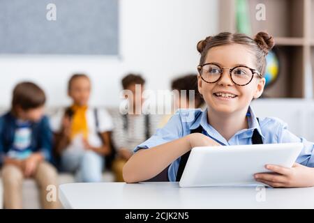 Selektive Fokus der Schülerin mit digitalen Tablet am Schreibtisch sitzen In der Nähe von Freunden im Hintergrund im Klassenzimmer Stockfoto