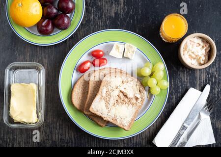 Mittags offenes Sandwich – zwei Scheiben Brot und Butter, Käse, Tomaten und Trauben auf einem Teller mit Obst, Krautsalat und einem Glas Orangensaft Stockfoto