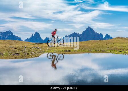 Nette Frau, die mit ihrem elektrischen Mountainbike die drei Zinnen Dolomiten fährt und sich im blauen Wasser eines kalten Bergsees spiegelt Stockfoto