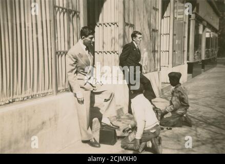 Antikes c1940-Foto, zwei junge Männer, die ihre Schuhe von afroamerikanischen Jungen auf einer Stadtstraße in den Schimmer bringen. Genaue Lage unbekannt, wahrscheinlich Neuengland, USA. QUELLE: ORIGINALFOTO Stockfoto